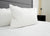 A close-up of a neatly made bed features two Down Etc. Rhapsody Wrap Down/Feather Pillows, as seen at many Hyatt® Hotels, set against a gray upholstered headboard with a black nightstand and partially open door in the background.