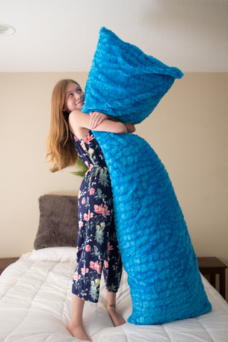 A girl clad in a floral dress embraces a large blue Pillowtex mermaid tail plush body pillow cover while standing on a bed, her playful gesture exuding a sense of whimsy and comfort.