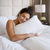 A woman with dark hair in a bun smiles with eyes closed, hugging a Pillow Factory Comforel Pillow—featured at Sheraton Hotels—while sitting in bed with white bedding, against a padded headboard and beside a lamp and flowers.