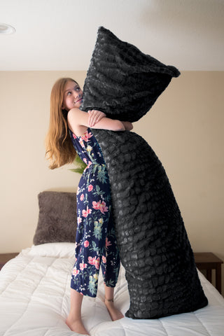 A young woman in a floral dress playfully holding onto a large, dark gray Pillowtex® body pillow cover almost her height while standing on a white-sheeted bed, with a warm, amused expression