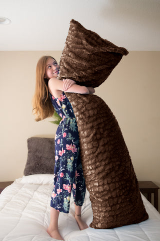 A joyful girl in a floral dress embraces a Pillowtex® Colorful Plush Faux Fur body pillow cover on a bed, her playful expression suggesting a moment of carefree happiness and imagination.