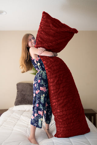 A joyful young girl in a floral dress playfully hugs a plush giant red Pillowtex® body pillow cover, standing on a white bed in a sunlit room, expressing delight and comfort in her cozy Pillowtex Colorful Plush Faux Fur.