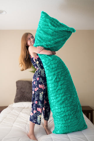 A young girl with long hair stands on a bed, happily hugging a large teal mermaid tail blanket, her pose mimicking that of a mermaid next to her plush Pillowtex® body pillow cover in colorful plush faux fur.