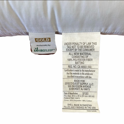 A close-up view of two fabric tags attached to the underside of a firm support pillow. The left tag is branded "Envirosleep Gold Pillow by Manchester Mills, exclusively made by Envirosleep® Gold Garneted Polyester Fiber Fill Pillow," and the right contains detailed care instructions and material composition.