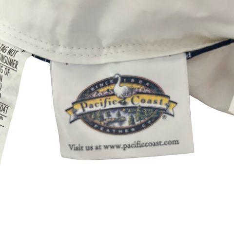 Close-up of a white fabric tag on a garment showing the logo of "Pacific Coast Feather Down Chamber Pillow," featuring a bird, plus a web address. The logo is colorful with text and.