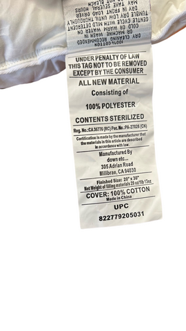 A close-up image of a law label on a Down Etc. Fairfax Firm Polyester Pillow, featuring printed text about material contents, certifications, and a barcode, with the bold headline "under penalty of law this tag not to