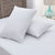 Two white Keeco Woodspring Ecoendure Pillows, as featured at many Choice® Hotels, rest on a quilted mattress—one upright by a wooden headboard, bathed in sunlight through sheer curtains and beside a blue glass lamp. Ideal for stomach sleepers.
