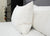 A close-up of two Down Etc. 25% White Goose Down / 75% White Goose Feather Pillows propped against a dark gray upholstered headboard on a bed with a white comforter, with a black dresser and stacked books partially visible nearby.