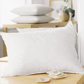 A neatly arranged bedroom setting with a crisp white pillow featuring Performa Fiberfill in the foreground and a stack of folded JS Fiber Quallofil Pillows in the background, accompanied by a small vase and decorative items.