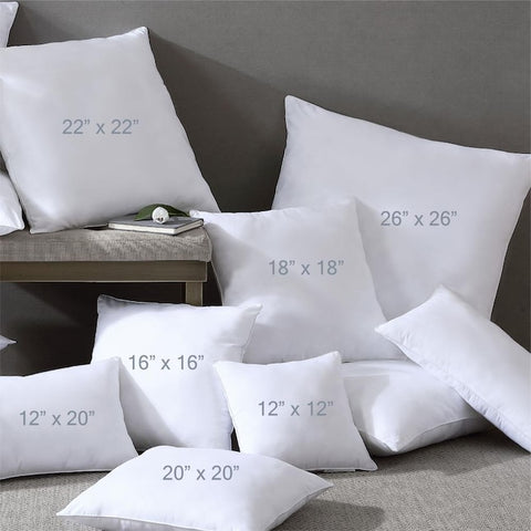 A collection of white Pillowtex Pillow Insert | White Duck Feather & Down, square, and rectangular duck feather pillow inserts of various sizes indicated by measurements, casually arranged against a gray backdrop, with a small end table holding a watch and glasses.