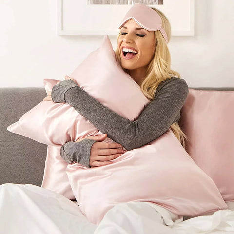 A joyful woman in a Pillows.com Mulberry Silk Sleep Eye Mask and comfy attire hugging a soft pillow while sitting on a bed with pink sheets, embodying relaxation and happiness.