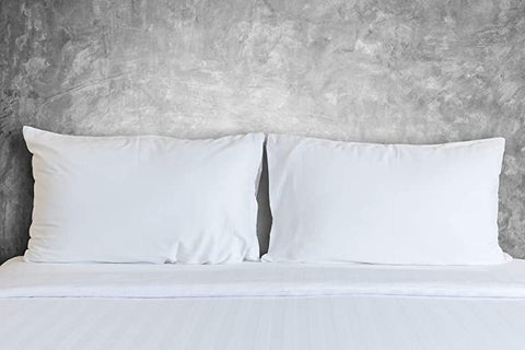 Two Encompass Group 50/50 White Goose Feather & Down Pillows sit neatly on a crisply made bed with white linens against a textured grey concrete wall, conveying a minimalist and contemporary bedroom aesthetic.