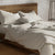 An unmade bed with crumpled Delilah Home GOTS certified, organic cotton sheets and pillows in a serene bedroom, featuring a wooden headboard and a soft glow from a lamp.