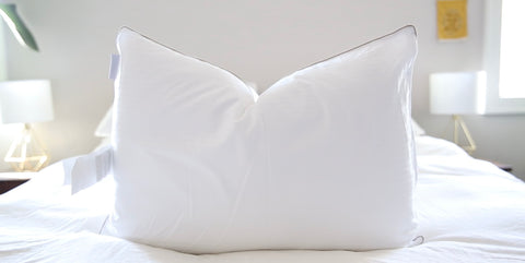 A pristine white Indulgence by Isotonic Synthetic Down Pillow from Carpenter sits at the center of a neatly made bed, flanked by two table lamps in a serene, well-lit bedroom setting.