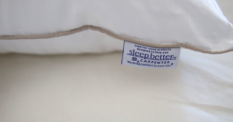 A close-up of a white pillow's corner showing a blue and white "sleep better" tag by Carpenter, suggesting a focus on comfort and sleep quality. This machine washable, down alternative Indulgence by Isotonic Synthetic Down Pillow.