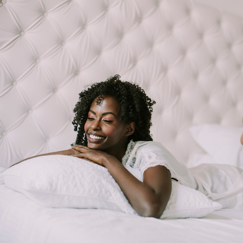 A young black woman laying on a white bed, surrounded by Pillowtex Down Alternative Soft Pillows.
