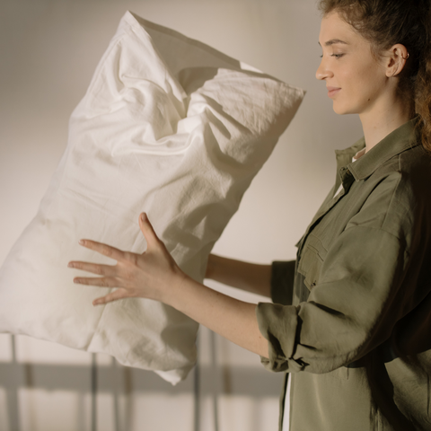A woman with curly hair in a green shirt holds a Restful Nights® Royal Loft® Polyester Pillow by Restful Nights against a soft beige background, seemingly arranging it, with a gentle and focused expression.