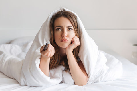 A pensive young woman lies in bed under a Suite Rest Recycled Polyester Blanket, remote in hand, with an expression of indecision or boredom.