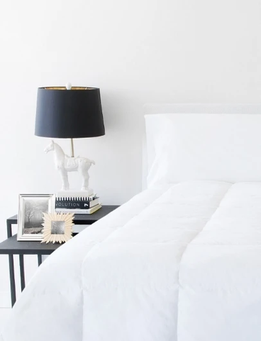 A bed with a white comforter and a lamp can be purchased using an electronic $100 gift certificate code from pillowsdotcom.