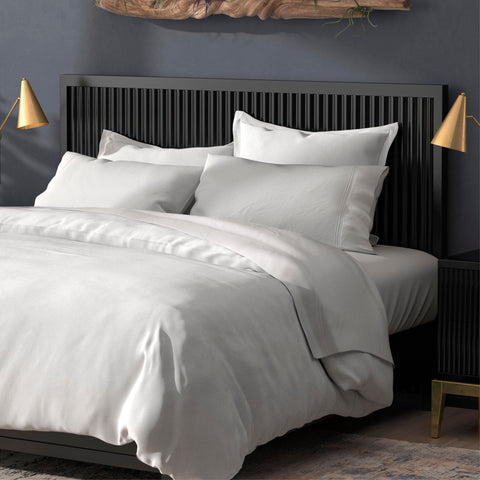 A modern bedroom featuring a neatly made bed with crisp white PureCare Pillow Sham Set | Soft Touch Bamboo bedding, complemented by a stylish, dark slatted headboard and matching side tables, accented by warm-toned.
