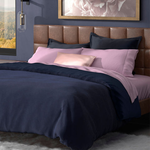 A neatly made bed with a plush leather headboard, featuring a combination of pink and navy-blue bedding with a PureCare Soft Touch Bamboo duvet cover and an artistic floral wall decor, set against a window with a