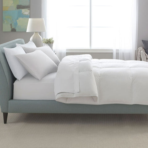 A neatly made bed with a Restful Nights down alternative comforter and crisp white bedding in a serene bedroom with soft blue and gray decor, natural light streaming through the window, and a modern abstract painting on the wall by Pacific Coast Feather Company.
