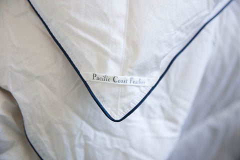 A close up of a white bed with a Pacific Coast Feather Company SuperLoft™ Down Comforter blue label.