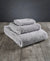 Three 100% Organic Cotton Bath Towels from Delilah Home, neatly stacked by size, rest on a rustic wooden surface. A smooth dark gradient background highlights their soft, fluffy texture and minimalist luxury.