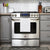 A modern stainless steel gas stove with a digital control panel, four knobs, and a Delilah Home 100% Organic Cotton Kitchen Towel hanging on the oven handle. Gray cabinets, kitchen tools, knife block, toaster, and white subway tile backsplash complete the space.
