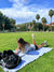 A young woman relaxes on a Delilah Home 100% Organic Cotton Sunset Beach Towel, reading on her tablet on a sunny campus lawn. She wears a gray sweatshirt and white sneakers, with palm trees and buildings in the background.