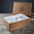An open brown box with Delilah Home’s logo displays a folded, light gray luxury hemp sheet from the Final Sale: Organic Hemp Bed Sheet Collection, wrapped in a white ribbon with small logos. The scene sits on wood against a dark background.