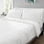 A neatly made bed features Delilah Home's Final Sale: Organic Hemp Bed Sheet Collection, with pillows and a duvet, in a sunlit bedroom with soft gray walls. A small nightstand with a plant and lamp sits beside the bed.