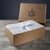 A brown Delilah Home gift box with a leaf and animal head logo sits open on gray wood, revealing pale gray Final Sale: Organic Hemp Bed Sheet Collection neatly folded and tied with white ribbon showing circular logos. Dark gray background.