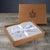 An open brown cardboard box with a leaf and seal logo reveals Delilah Home's Final Sale: Organic Hemp Bed Sheet Collection. The light grey sheets are neatly folded and tied with a white satin ribbon featuring a circular logo bow.