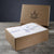 A brown Delilah Home box with a leaf and animal logo sits open on wood, displaying the Final Sale: Organic Hemp Bed Sheet Collection wrapped in white tissue and tied with a ribbon adorned with small circular logos.