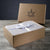 An open Delilah Home box with a leaf and fist logo sits on a gray wood surface. Inside, Final Sale: Organic Hemp Bed Sheet Collection is neatly folded and tied with a white ribbon featuring circular emblems. The logo appears on the lid behind.