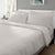 A neatly made bed features Delilah Home’s Final Sale: Organic Hemp Bed Sheet Collection in light gray, paired with matching pillowcases. Sunlight streams in, highlighting a serene space with a side table, plant, and framed abstract art.