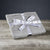A neatly folded light gray Delilah Home Final Sale: Organic Hemp Bed Sheet Collection, tied with a white ribbon featuring circular logos and a central bow, rests on wood with a dark gray backdrop—luxury comfort and style in organic hemp bedding.