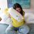 A woman in a yellow sweater and jeans sits cross-legged on a bed, happily hugging an Envirosleep Dream Surrender Polyester Pillow by Manchester Mills with labels attached. A colorful abstract painting with blue and green hues adorns the wall above a bed covered by a blue-gray coverlet.