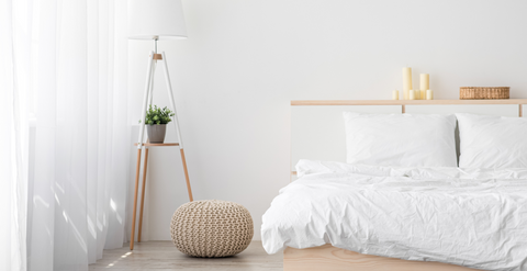 A minimalist bedroom with a white bed featuring a Restful Nights Eco Endure Pillow | Polyester Medium-Firm, crumpled duvet, adjacent wooden floor lamp, and a knitted pouf. A plant and candles on