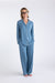 A light-haired woman with glasses stands barefoot on a white background, smiling. She wears the Sleep Again Pillows Long Sleeve Bamboo Pajama Set—light blue, loose-fitting, with white piping and buttons—breathable and perfect for relaxing.