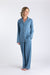 A blonde woman in a loose updo smiles to her left, wearing the Sleep Again Pillows Long Sleeve Bamboo Pajama Set—light blue with white piping and matching wide-leg pants—breathable sleepwear ideal for relaxing barefoot on a white background.