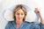 A woman rests with eyes closed, head supported by the Sleep Again Pillows Cervical Memory Foam Pillow for Neck & Shoulder Support. She appears relaxed in blue pajamas, accessorized with layered gold necklaces and rings.