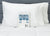 Two white pillows with Pillowtex Cotton Pillowcase Set rest against a grey headboard. In front, the blue and white packaging from Pillowtex, highlighting features and warnings, sits on a white mattress.