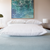 The bed has a soft, white hypoallergenic pillow centered on a light blue quilted blanket, with two Manchester Mills Envirosleep® Dream Surrender Firm pillows, ideal for side sleepers. A framed abstract artwork featuring blue and green hues decorates the background.