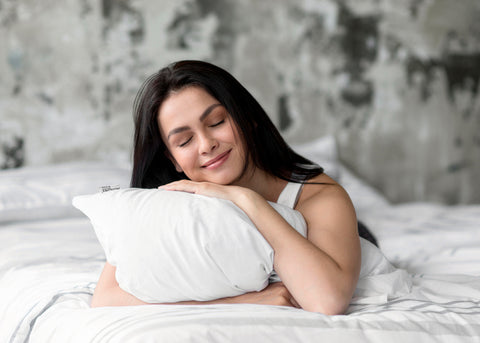 A contented woman with long dark hair, clad in a white tank top, gently smiles while embracing a soft Pillowtex White Goose Down & Feather Pillow, enjoying a peaceful moment in a serene, gray-toned bedroom setting.