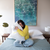 A woman in a yellow sweater and jeans sits on a bed with a light blue cover, hugging an Envirosleep Dream Surrender Polyester Pillow by Manchester Mills. Medium support pillows accompany her. Beside her are two tables with lamps under a large abstract artwork, and she looks content.