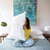 A person in a bright yellow sweater and light blue jeans sits on a bed with teal blanket and white pillows, holding the Manchester Mills Envirosleep Dream Surrender Polyester Pillow for medium support. Geometric lamps flank the bed, beneath a large painting resembling water.