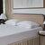 A neatly made bed with a beige upholstered headboard features two La-Z-Boy Down Alternative Pillows from La-Z-Boy in white patterned pillowcases and a white textured mattress cover, complemented by beige curtains, bedside tables with white lamps, and abstract artwork.