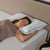 A woman with brown hair sleeps on a white mattress, resting her head on a La-Z-Boy Down Alternative Pillow with black piping. She wears a sleeveless taupe top, arm bent by her head, against a beige headboard.
